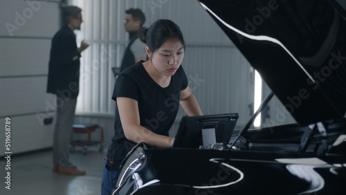 An asian female mechanic in uniform in a car service inspecting and checking the engine in a car with tablet, while a worker talking to a client