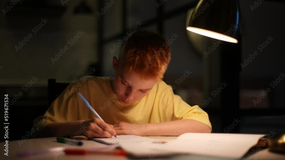 Thoughtful pupil boy studying at home writing in notebook with pen sitting at table under light of lamp. Caucasian schoolboy doing homework in dark room at evening. Shooting in slow motion. Thoughtful pupil boy studying at home writing in notebook with pen sitting at table under light of lamp. Caucasian schoolboy doing homework in dark room at evening. Shooting in slow motion.