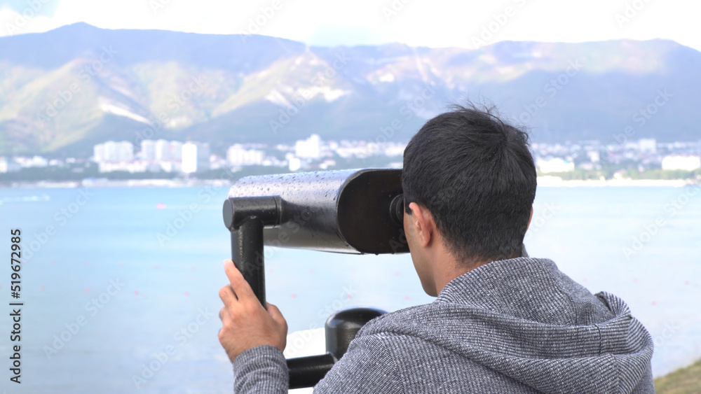A man looking through coin binoculars on fantastic mountain and sea view. Media. Male tourist looks at the coastal city through coin binoculars.