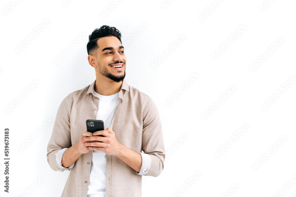 Positive attractive indian or arabian guy, holding smartphone in hand, chatting online, browsing internet, looking to the side, thinking, dreaming, standing on isolated white background, smiling Positive attractive indian or arabian guy, holding smartphone in hand, chatting online, browsing internet, looking to the side, thinking, dreaming, standing on isolated white background, smiling