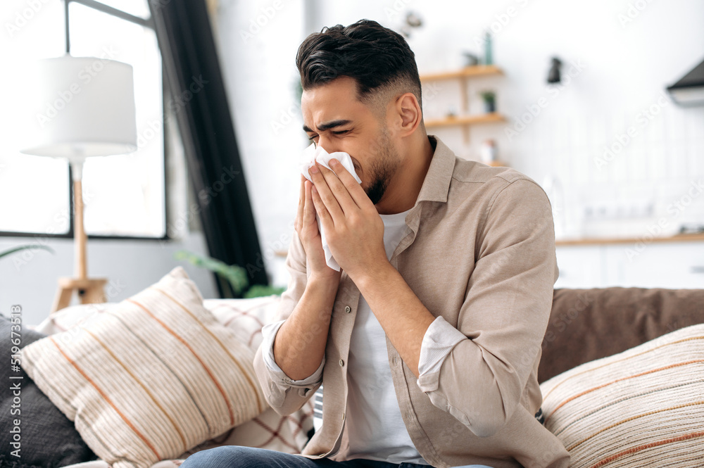 The concept of illness, runny nose. Unhealthy sad arabian or indian man sits at home in the living room on the couch, feels unwell, suffers from a runny nose, sneezing, uses napkins, needs treatment The concept of illness, runny nose. Unhealthy sad arabian or indian man sits at home in the living room on the couch, feels unwell, suffers from a runny nose, sneezing, uses napkins, needs treatment