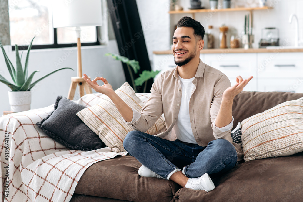 Happy positive calm relaxed indian or arabian man in casual clothes, sitting alone at home in living room on the couch and meditating in the lotus position, take care of his mental health, smiling