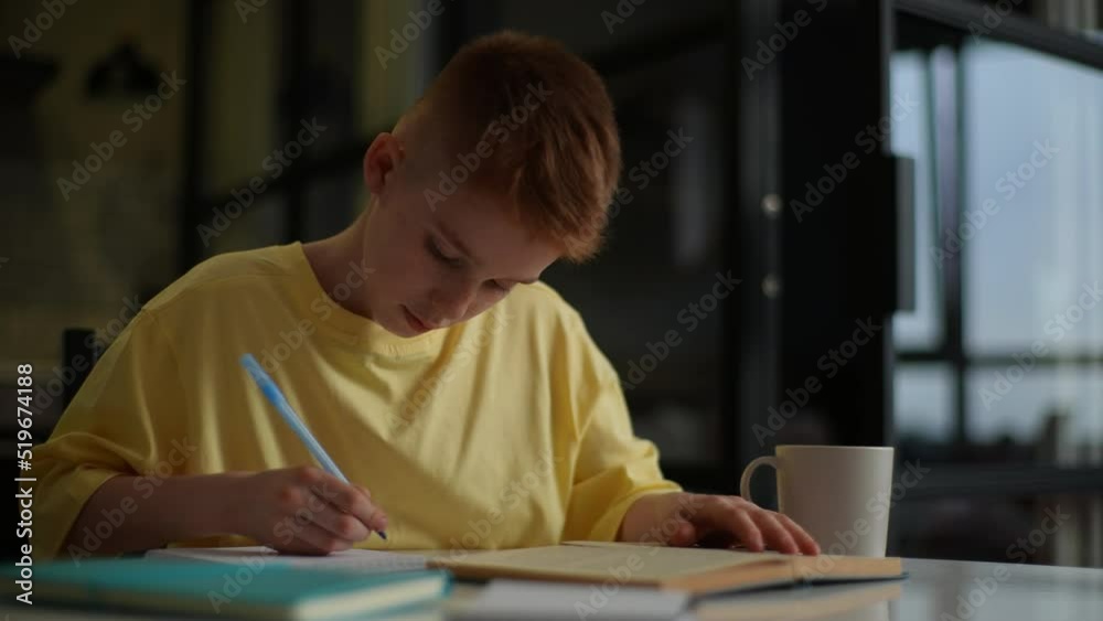 Cute red-haired pupil boy studying at home writing in exercise book doing homework, learning at home table. Smart schoolboy with freckles studying, reading textbook, flipping page of paper book. Cute red-haired pupil boy studying at home writing in exercise book doing homework, learning at home table. Smart schoolboy with freckles studying, reading textbook, flipping page of paper book.