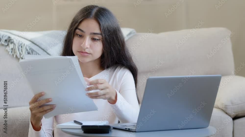 Focused young freelancer housewife arabian woman girl holds papers utility bills makes remote payment e-pay through e-bank app on laptop texting on keyboard data analysis studying alone in living room