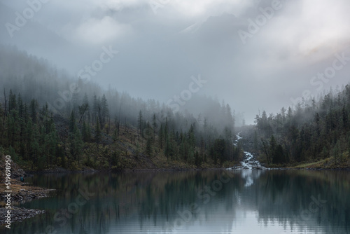 Mountain creek flows from forest hills into glacial lake in mysterious fog. Small river and coniferous trees reflected in calm alpine lake in early morning. Tranquil misty scenery with mountain lake. © Daniil