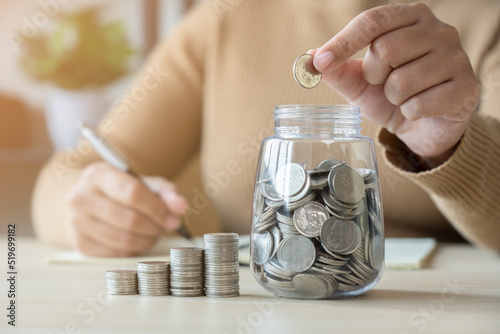 Woman with coins jar taking note to checklist expenses. Concept of doing accounting plan with coin jar and woman writing in a book.