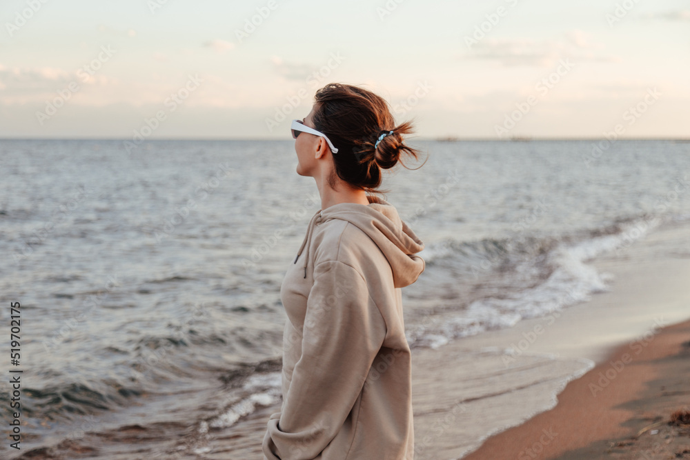 Woman standing by the lake shore in hoodie and glasses alone looking at waves Woman standing by the lake shore in hoodie and glasses alone looking at waves
