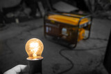 A transparent incandescent lamp glows brightly close-up against the background of a yellow gasoline electric generator in the dark