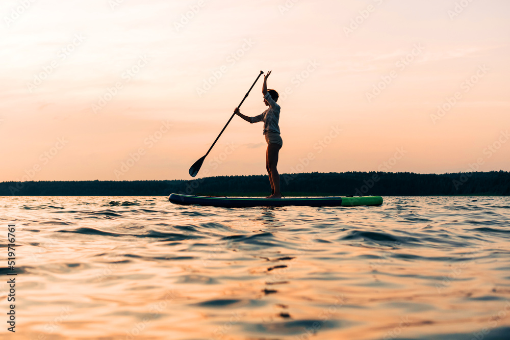 Joyful sport woman is training on a SUP board on a large lake during the evening. Stand up paddle boarding - awesome active recreation in nature.