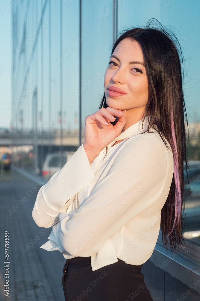Young woman with dark long hair and plump pink lips smiles posing against blurry city in windy weather. Stylish lady wears formal business clothes closeup