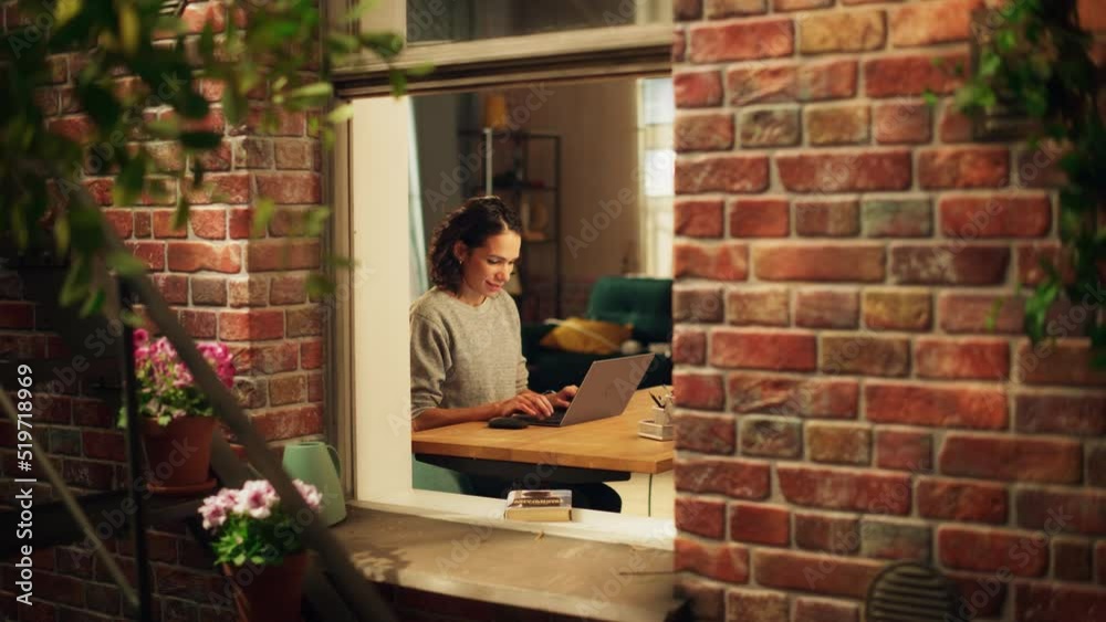 Hispanic Woman Using Laptop Computer, Sitting in Living Room im Her Apartment. Professional Female Working Remotely Online, Planning, Brainstorming New Project. Shot Into the Window From Outside. Hispanic Woman Using Laptop Computer, Sitting in Living Room im Her Apartment. Professional Female Working Remotely Online, Planning, Brainstorming New Project. Shot Into the Window From Outside.