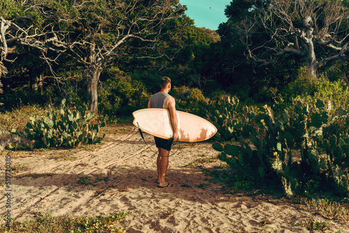 Mężczyzna surfer z deską surfingową, idący wybrzeżem na tle jungle, aktywny wypoczynek. © insomniafoto