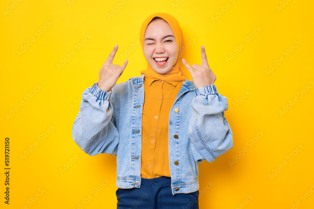 Cheerful young Asian woman in jeans jacket showing rock and roll gesture isolated over yellow background Cheerful young Asian woman in jeans jacket showing rock and roll gesture isolated over yellow background