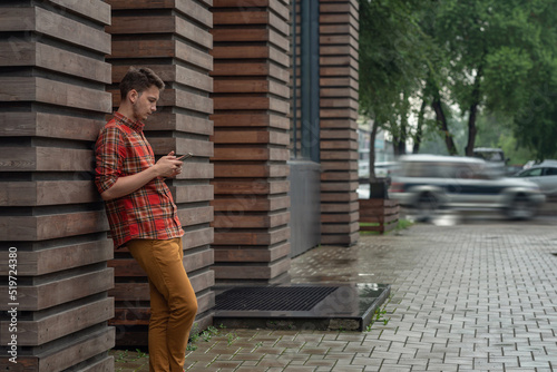 a young man in a red plaid shirt stands leaning against the wall and looks at the phone