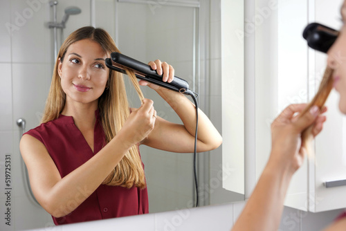 Girl using steam straightener to style hair at the mirror on bathroom photo