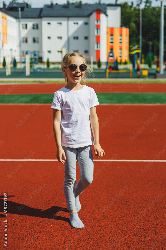 A little blonde girl is running on the sports field and smiling, she is wearing sunglasses and a white T-shirt. Summer holidays and vacation time.
