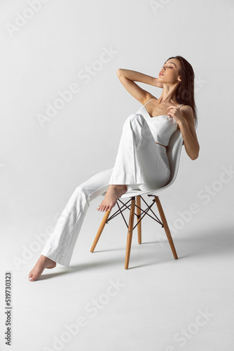 The full length image of a young brunette woman with makeup and hairstyle, dressed in white, sits fashioable on the chair.