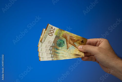 A spread of 200 Swiss Francs banknotes, held in hand by a Caucasian male. Close up studio shot, isolated on blue background