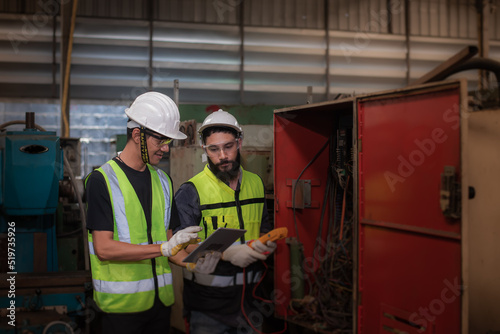 Young electrician latin mixed-race and asian engineer in safety uniforms workwear Inspecting and maintaining the machine's electrical control cabinet in heavy industrial manufacturer factory.