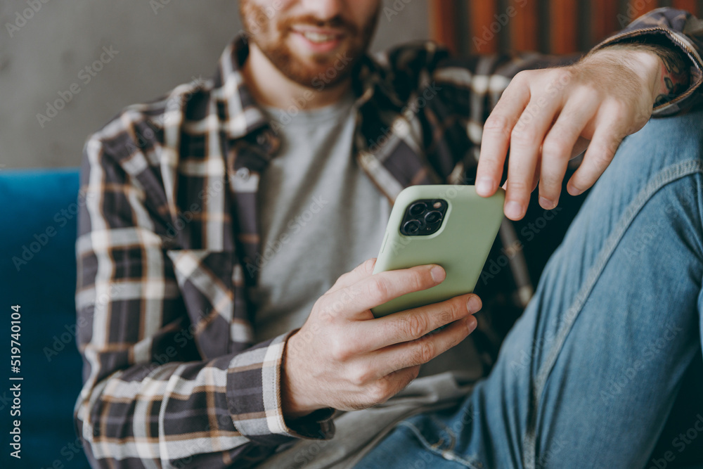 Cropped young caucasian man 20s he wears brown shirt hold in hand use mobile cell phone sitting on blue sofa in own living room apartment stay home indoor flat on weekends. People lifestyle concept. Cropped young caucasian man 20s he wears brown shirt hold in hand use mobile cell phone sitting on blue sofa in own living room apartment stay home indoor flat on weekends. People lifestyle concept.