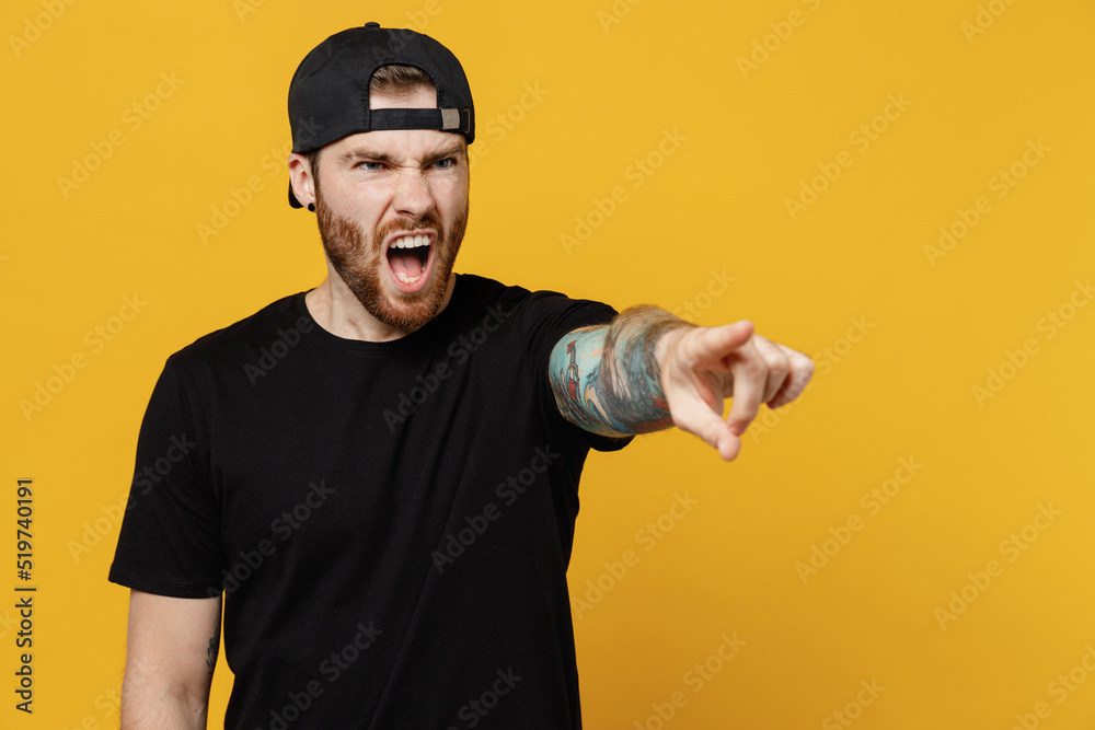 Young sad indignant angry bearded tattooed man 20s he wears casual black t-shirt cap point index finger aside scream isolated on plain yellow wall background studio portrait. People lifestyle concept. Young sad indignant angry bearded tattooed man 20s he wears casual black t-shirt cap point index finger aside scream isolated on plain yellow wall background studio portrait. People lifestyle concept.