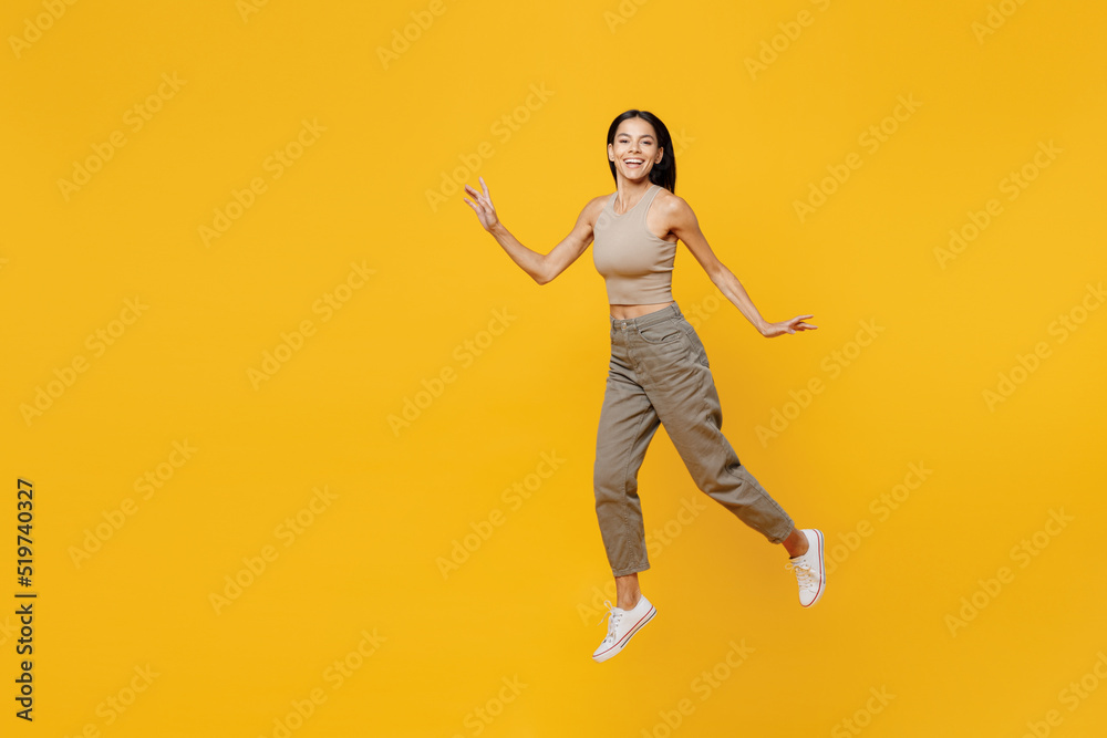Full body young satisfied cheerful smiling happy latin woman 30s she wearing basic beige tank shirt jump high like flying isolated on plain yellow backround studio portrait. People lifestyle concept.