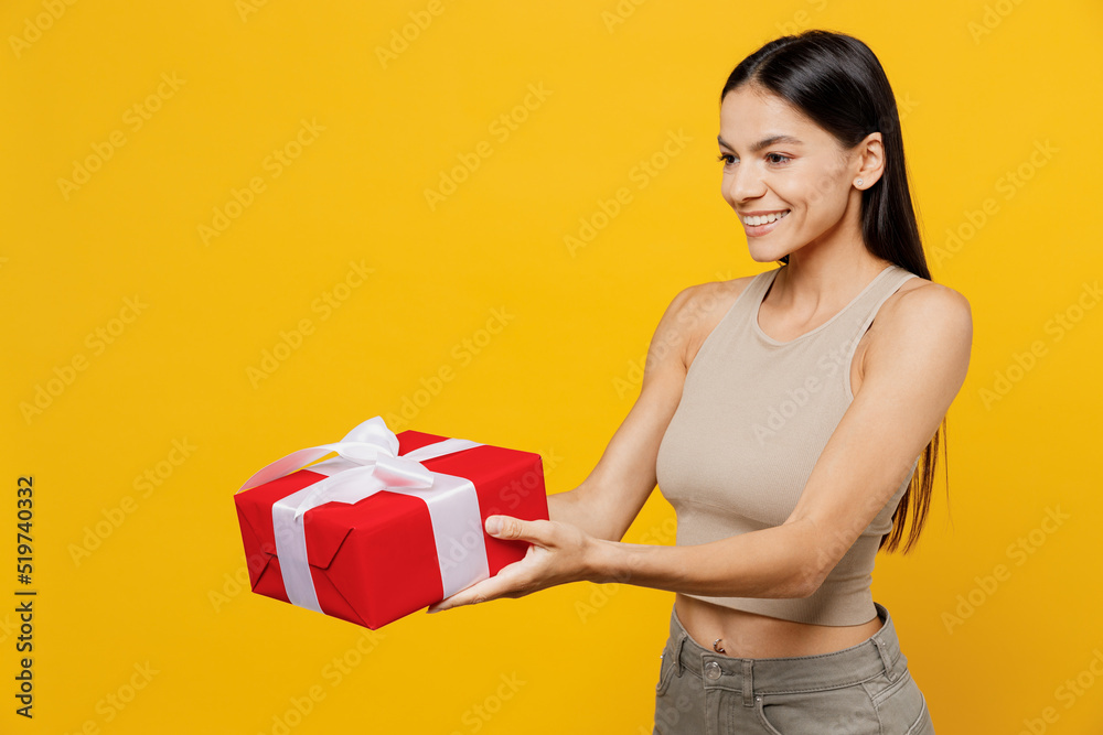 Side view young smiling happy cheerful cun cool latin woman 30s she wear basic beige tank shirt hold in hand give red present box with gift ribbon bow isolated on plain yellow color backround studio. Side view young smiling happy cheerful cun cool latin woman 30s she wear basic beige tank shirt hold in hand give red present box with gift ribbon bow isolated on plain yellow color backround studio.