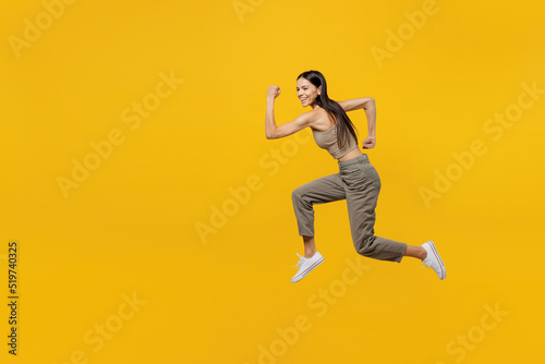 Full body side view smiling happy fun cool young latin woman 30s she wear beige tank shirt jump high run fast look camera isolated on plain yellow backround studio portrait. People lifestyle concept.