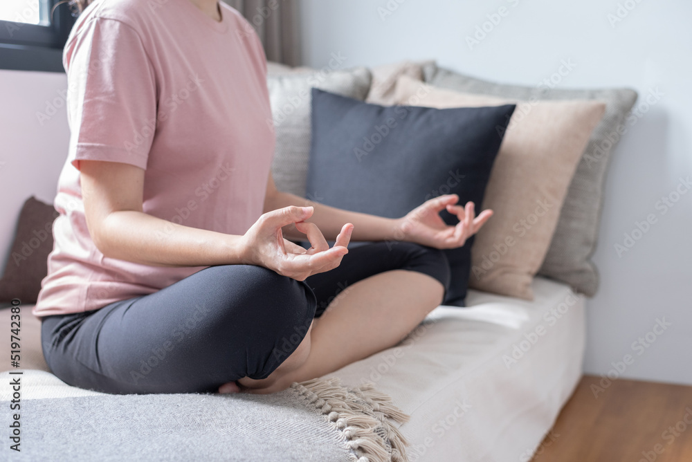 Close up, side view with copy space of unrecognizable woman sitting in Ardha Padmasana position on couch near windows, practicing yoga meditation, doing half Lotus pose with mudra gesture.