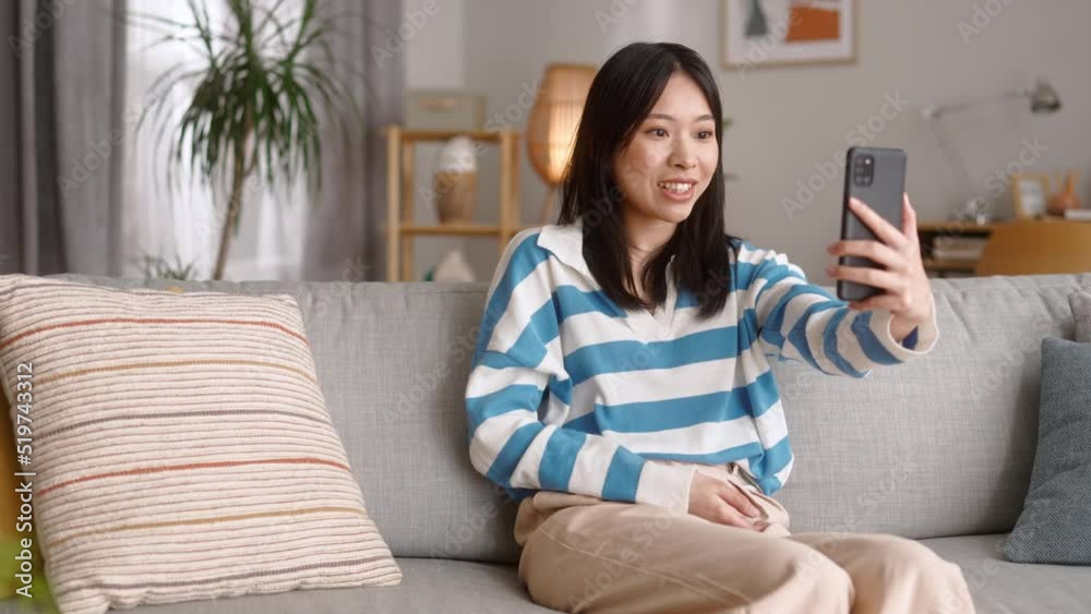 Smiling asian woman sitting on couch at home and making hello gesture to camera on modern smartphone. Young female having video call with friend online. Smiling asian woman sitting on couch at home and making hello gesture to camera on modern smartphone. Young female having video call with friend online.