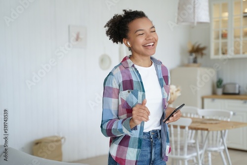 Excited happy girl in airpods is holding mobile phone. Teenage girl is singing in weekend morning. photo