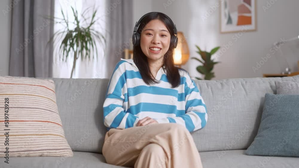 Portrait of young smiling woman in wireless headphones sitting on comfy sofa in bright apartment. Beautiful asian female in casual wear looking at camera and listening music. Portrait of young smiling woman in wireless headphones sitting on comfy sofa in bright apartment. Beautiful asian female in casual wear looking at camera and listening music.
