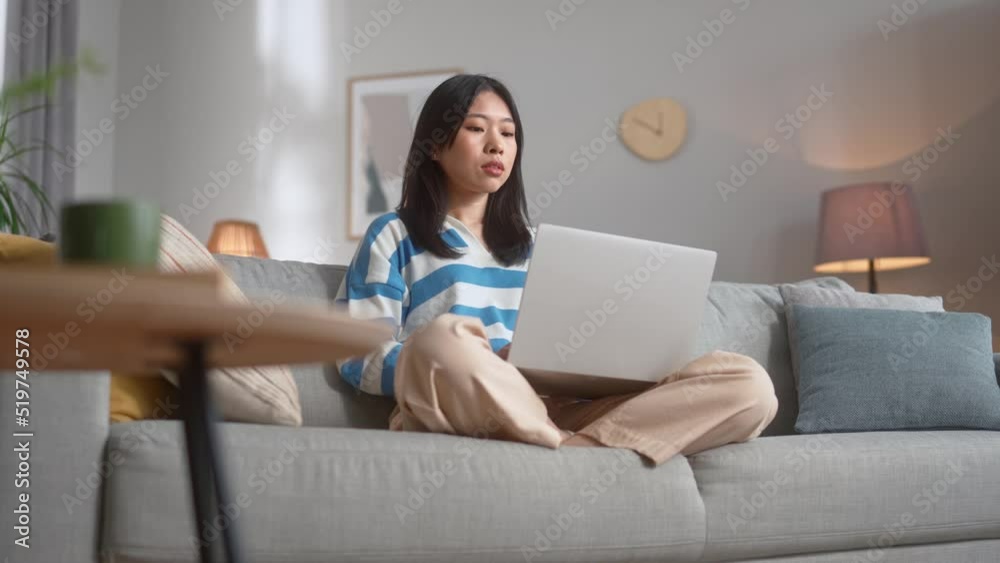 Happy young lady in stylish outfit surfing internet on wireless laptop while sitting on cozy sofa. Pretty chinese woman staying at home and using modern gadget. Happy young lady in stylish outfit surfing internet on wireless laptop while sitting on cozy sofa. Pretty chinese woman staying at home and using modern gadget.