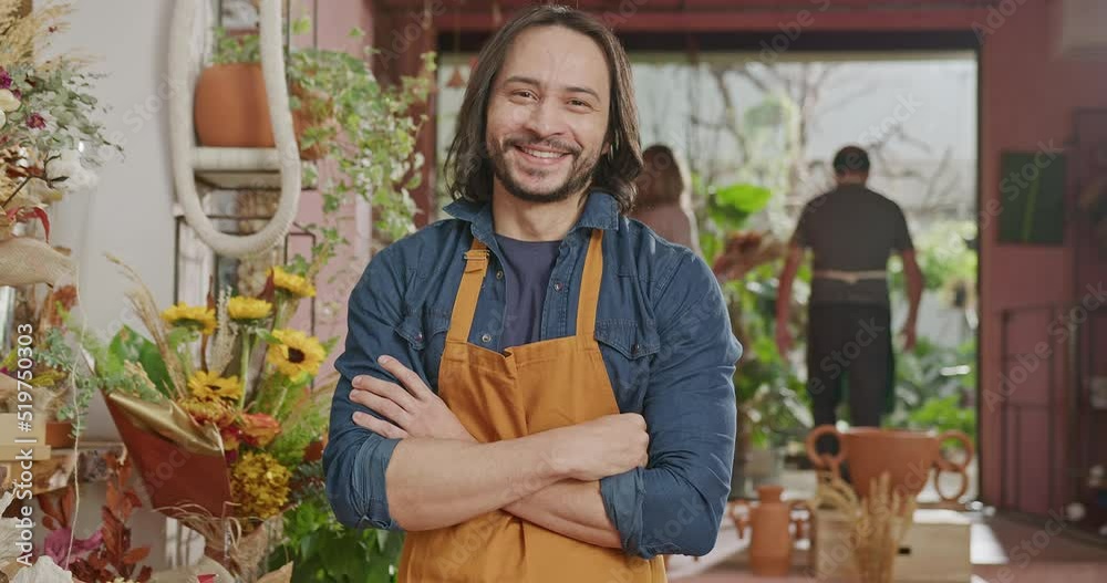 Portrait of a young man wearing apron standing inside small business flower shop with people working in the background. A happy male staff looking at camera Portrait of a young man wearing apron standing inside small business flower shop with people working in the background. A happy male staff looking at camera