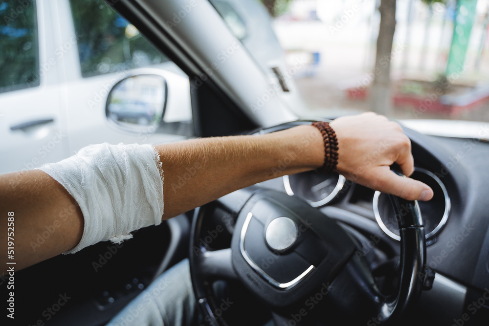 Naklejka premium The guy sits behind the wheel of the car, the injured hand lies on the steering wheel, the elbow hurts, the joint is bandaged, the man's hand is in the car.