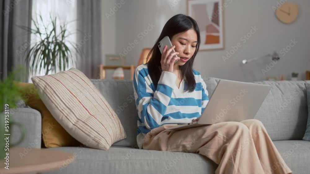 Charming chinese woman in casual clothes sitting on couch with laptop on knees and talking on modern smartphone. Concept of people, technology and lifestyles. Charming chinese woman in casual clothes sitting on couch with laptop on knees and talking on modern smartphone. Concept of people, technology and lifestyles.