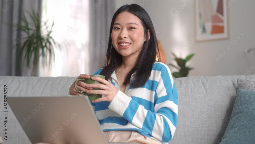 Portrait of young asian woman sitting on comfortable couch with laptop on knees at home. Attractive happy female holding cup of coffee and looking at camera indoors. Portrait of young asian woman sitting on comfortable couch with laptop on knees at home. Attractive happy female holding cup of coffee and looking at camera indoors.