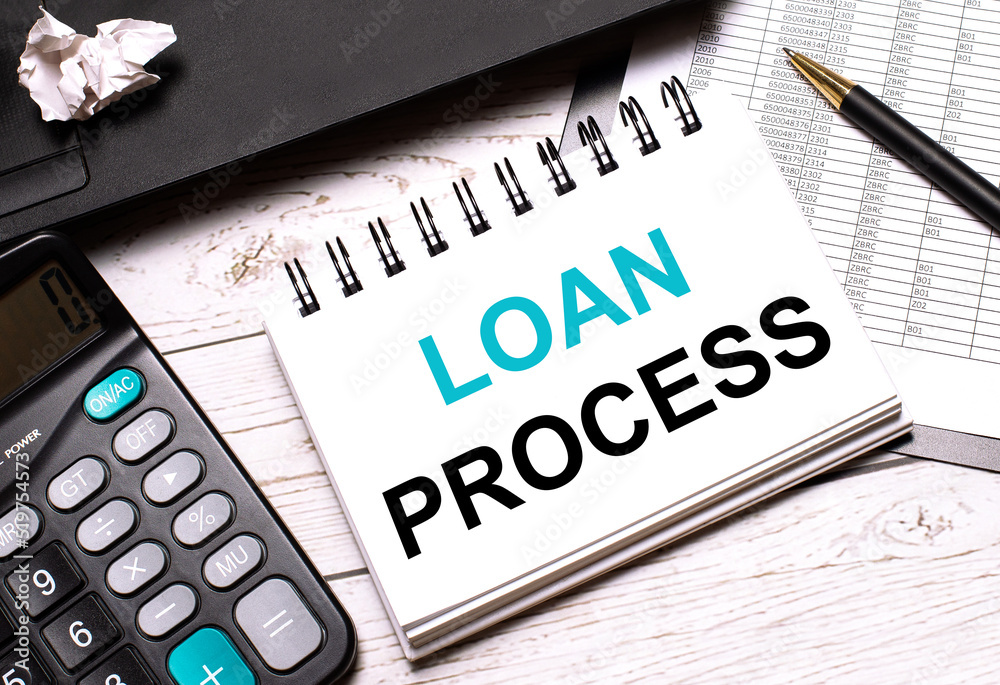 On a light-colored office table is a computer, a black calculator, a pen, and a notebook with the text LOAN PROCESS. Business concept. On a light-colored office table is a computer, a black calculator, a pen, and a notebook with the text LOAN PROCESS. Business concept.