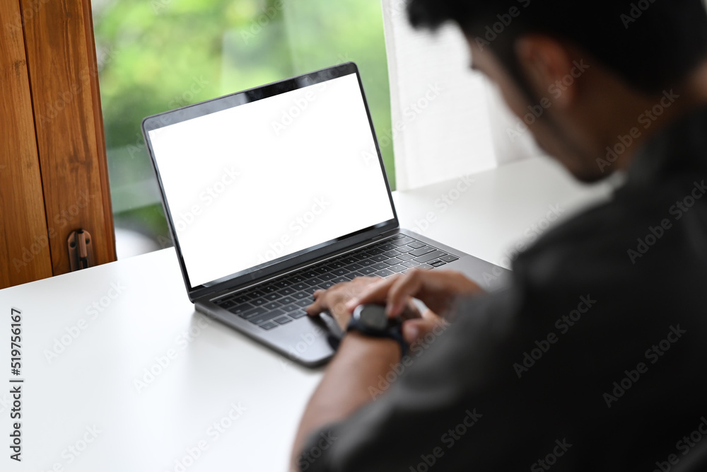 Young male employee sitting front of laptop computer at office desk and checking time in smart watch. Over shoulder view Young male employee sitting front of laptop computer at office desk and checking time in smart watch. Over shoulder view
