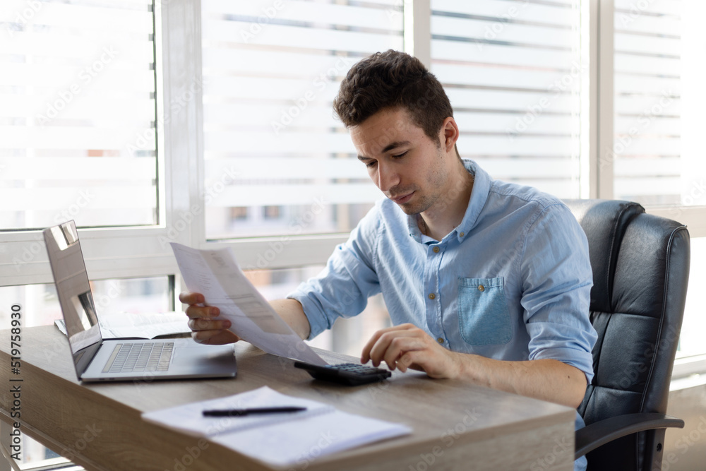 Young businessman checking bills, holding receipt, using calculator, accountant calculating company expenses, financial report, sitting at wooden work desk, young male planning budget Young businessman checking bills, holding receipt, using calculator, accountant calculating company expenses, financial report, sitting at wooden work desk, young male planning budget