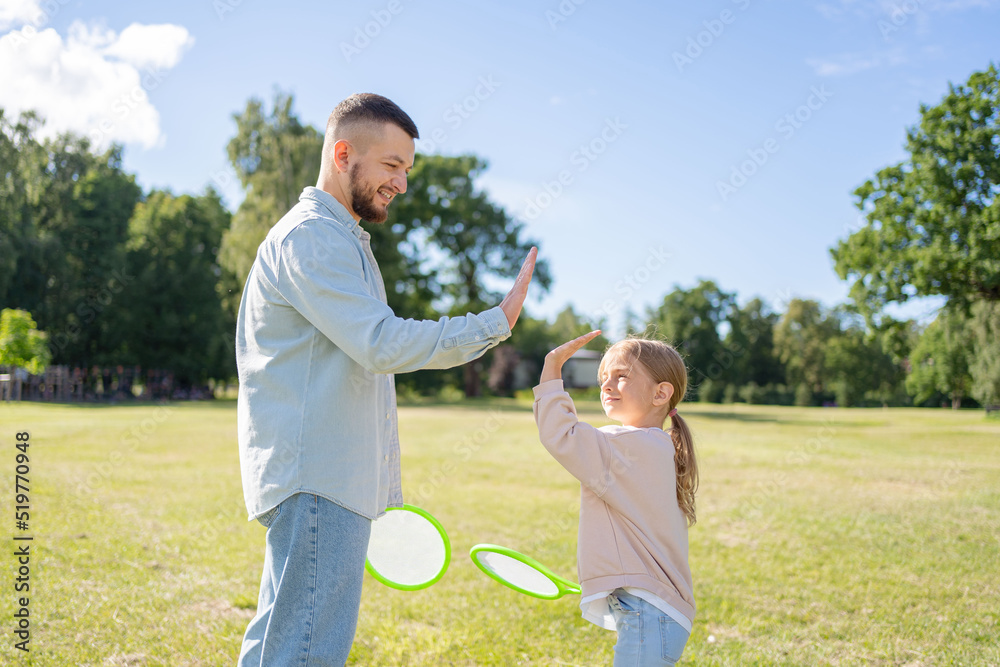 Fototapeta premium Cute little girl with her father with badminton rackets outdoors. Young man with daughter playing badminton together in park. Active lifestyle.