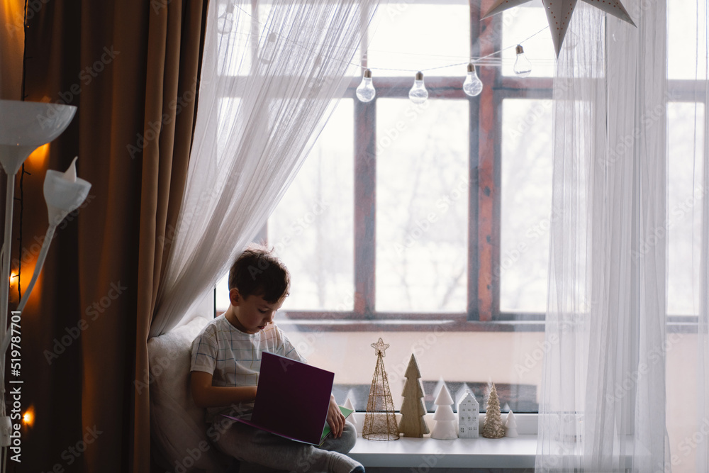 Cute boy is reading a book sitting on the windowsill near the window. Cozy home with decorative Christmas trees. Sweet home. Winter holidays lifestyle.