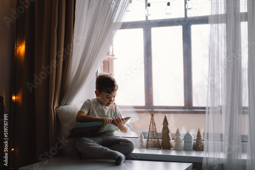 Cute boy is reading a book sitting on the windowsill near the window. Cozy home with decorative Christmas trees. Sweet home. Winter holidays lifestyle. Free Download Cute boy is reading a book sitting on the windowsill near the window. Cozy home with decorative Christmas trees. Sweet home. Winter holidays lifestyle. - GFXLoad