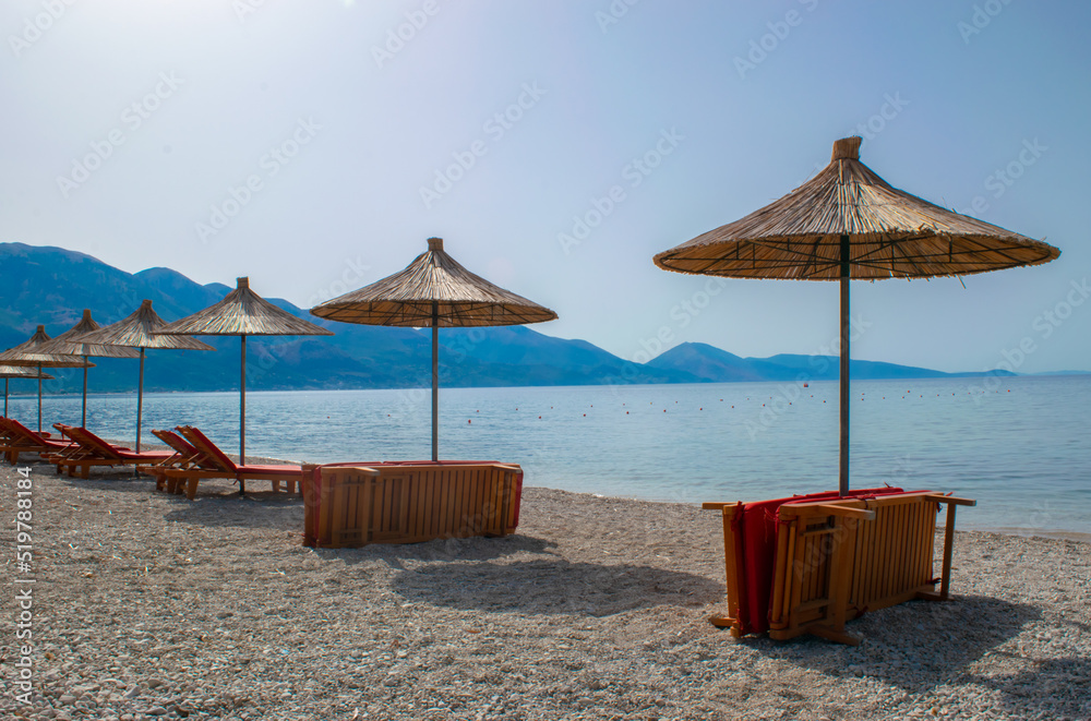 Beach umbrellas, and sun loungers on pebble beach. Qeparo village. Albania. Ionian Sea. Vacation sea concept. Summer sunny seaside landscape.