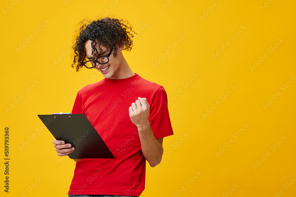 Joyful happy myopic young student man in red t-shirt funny eyewear holds tablet folder with study notes posing isolated on over yellow studio background. Free place for ad. Education College concept Joyful happy myopic young student man in red t-shirt funny eyewear holds tablet folder with study notes posing isolated on over yellow studio background. Free place for ad. Education College concept