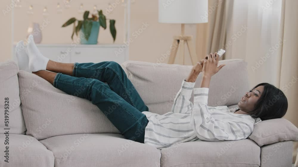 Relaxed carefree young calm woman lying on comfortable couch in cozy living room typing message on smartphone chatting online checks email communicates on dating site resting alone at home relaxes Relaxed carefree young calm woman lying on comfortable couch in cozy living room typing message on smartphone chatting online checks email communicates on dating site resting alone at home relaxes