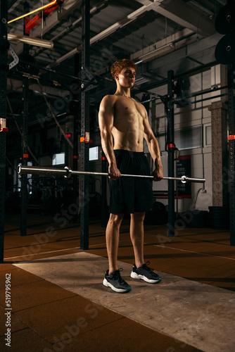 Portrait of shirtless sportive young man training, lifting barbell, doing exercises isolated over gym background