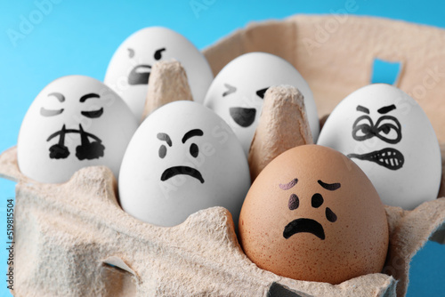 Brown egg with upset face among aggressively disposed white ones in carton box on turquoise background, closeup. Bullying concept