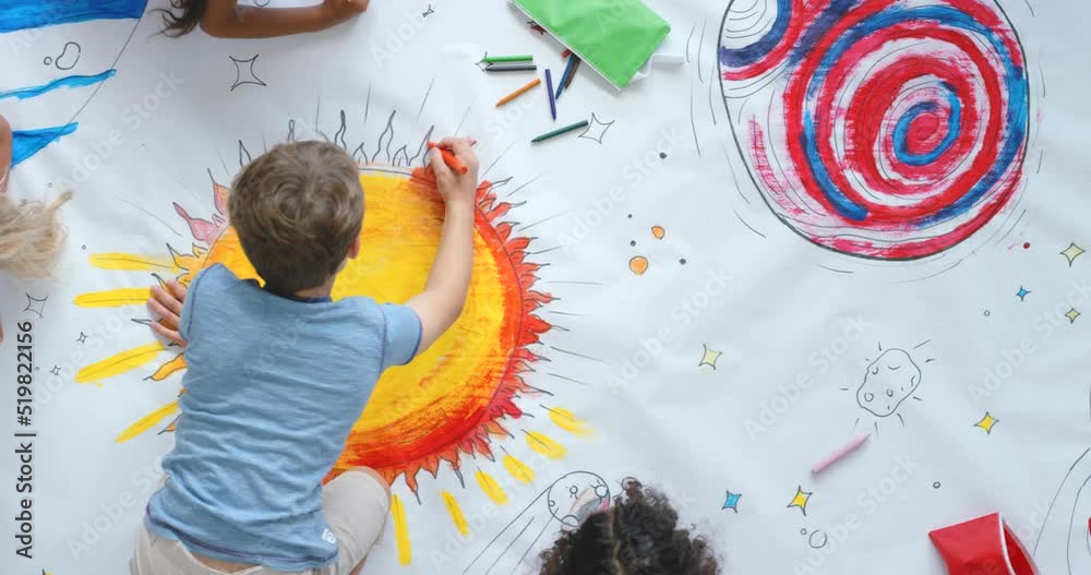 Young children and friends paint colorful space pictures indoors on the floor in kindergarten from the above view. Kids being creative painting science fiction drawings with colors on a big sheet.