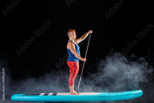 A woman with a mohawk on a sub board with an oar in the fog on a black background.